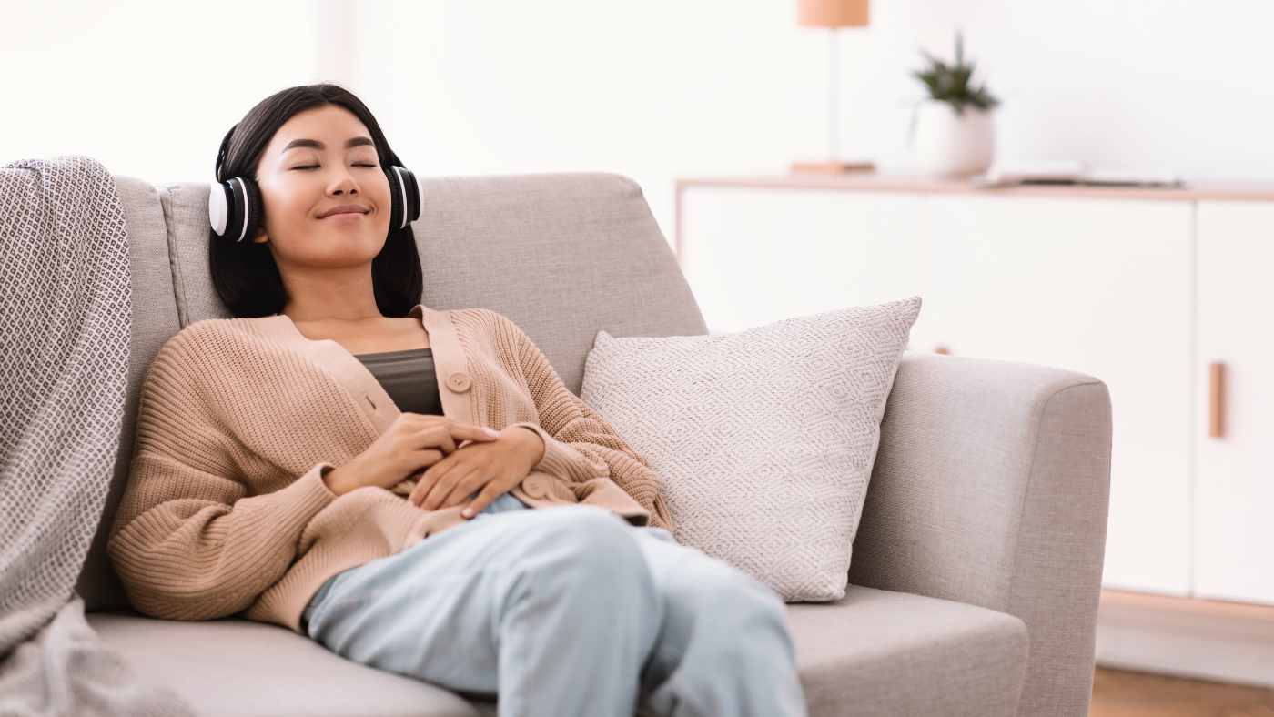 Asian woman with tan open cardigan and jeans with headphones on listening to music on a comfy couch to help with sleep