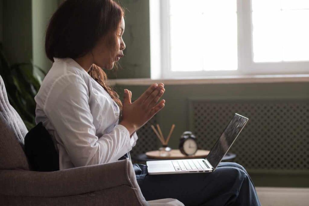 Black woman with white shirt holding laptop on her lap accessing online teletherapy now that she has the answer to the question does aetna cover mental health servcies