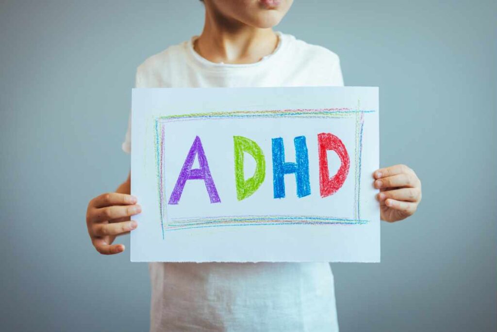 Child holding up a sign that reads ADHD. The letters are in all different colors and the child is wearing a white t-shirt.