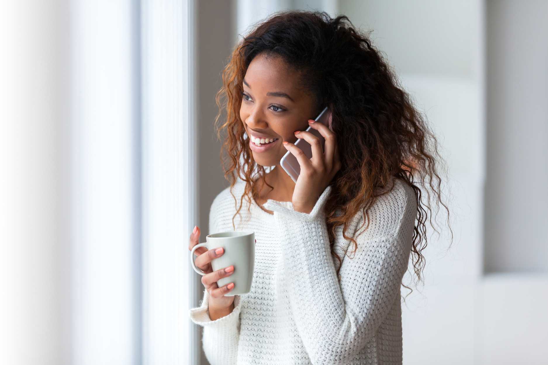 Happy Black woman on the phone with Rowan Center for Behavioral Medicine Scheduling her consult while holding a coffee. She's wearing a white sweater.