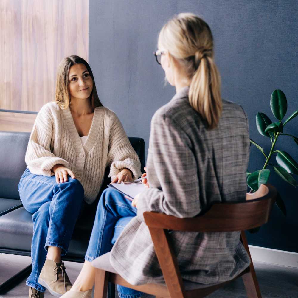 Patient with blue jeans and a tan cardigan sitting on a couch working with a licensed psychologist wearing plaid jacket for anxiety treatment 1 scaled for web. Both are sitting in a psychologist's office.