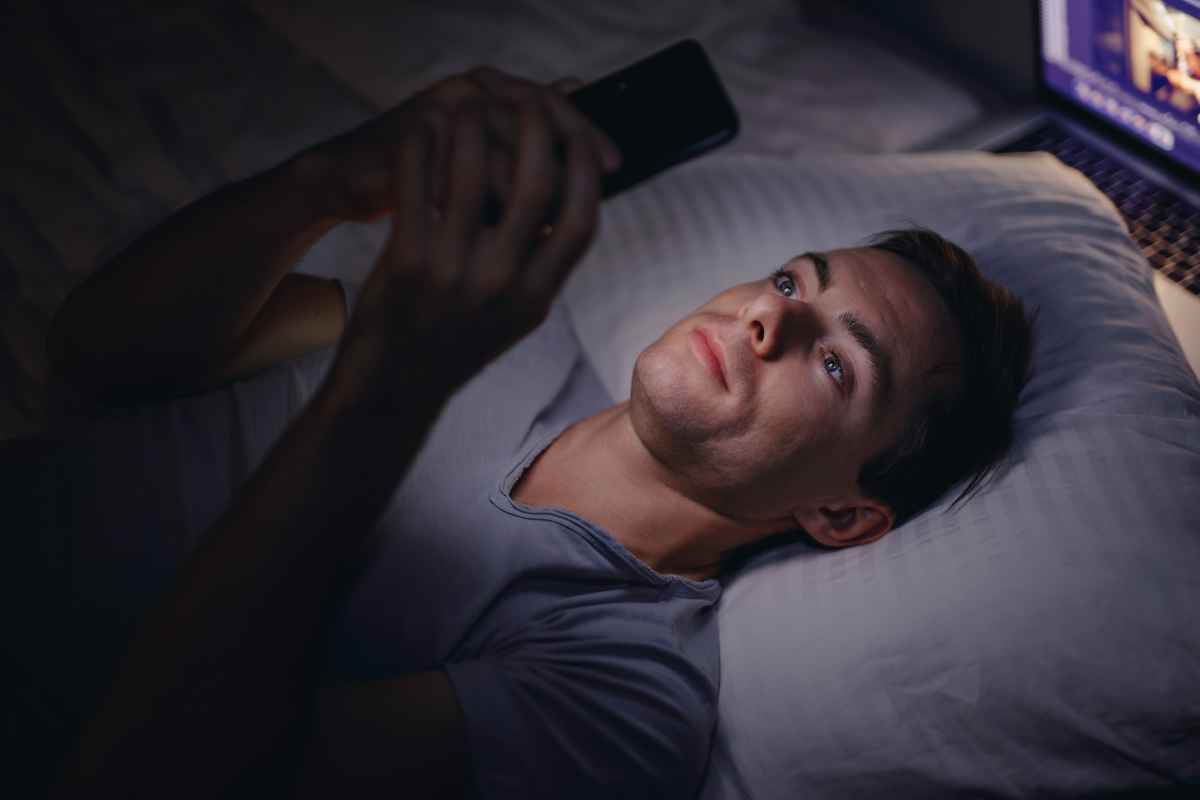 Sleep Pathophysiology How Screen Time Before Bed Ruins Sleep Featured Image - Man in bed laying on a pillow with the light of the phone lighting up his face