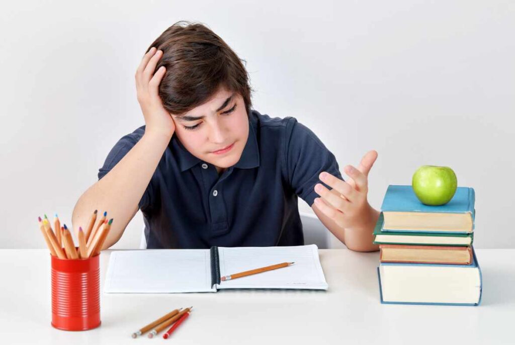 Student struggling with ADHD at their desk staring at paper and pencil with a look of clear frustration. For giggles, there is a green apple on top of books to the left of the student clearly identifying they are at school