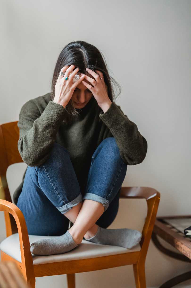 Woman with hair in her face and hands pushing through her hair experiencing anxiety.  She has on a green shirt and blue pants and is sitting on a chair with legs crossed.